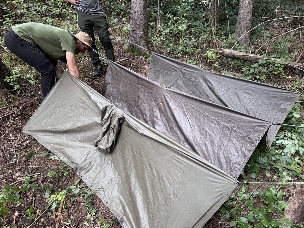 Zwei Teilnehmer spannen gemeinsam eine Tarp-Unterkunft im Wald auf